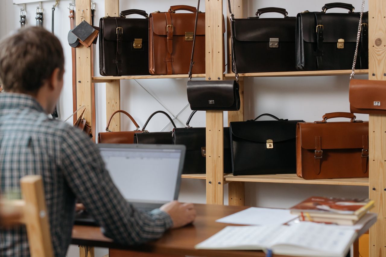 Services Man at desk using laptop near shelves filled with leather bags.